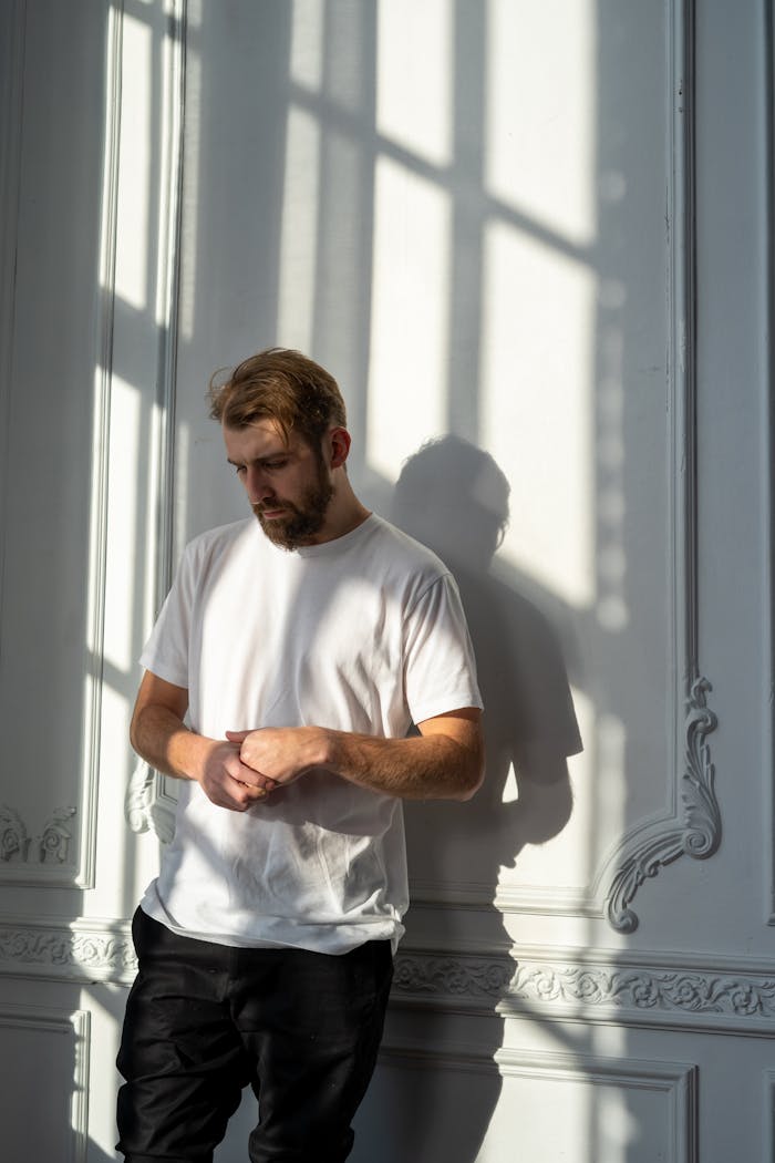 A thoughtful bearded man in a white t-shirt stands in a sunlit room with ornate walls.